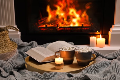 Cups of hot drink, candle and open book near fireplace indoors Photo of Cups of hot drink, candle and open book near fireplace indoors