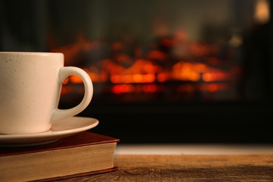 Cup with hot drink and book on table against fireplace, space for text Photo of Cup with hot drink and book on table against fireplace, space for text