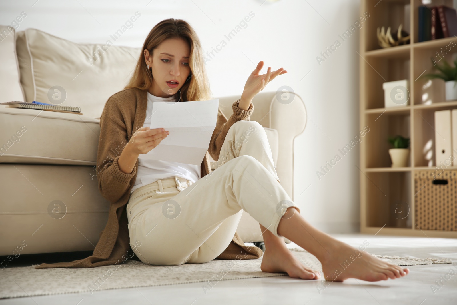 Shocked woman reading letter while sitting on floor near sofa at home Photo of Shocked woman reading letter while sitting on floor near sofa at home