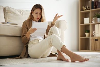 Shocked woman reading letter while sitting on floor near sofa at home Photo of Shocked woman reading letter while sitting on floor near sofa at home
