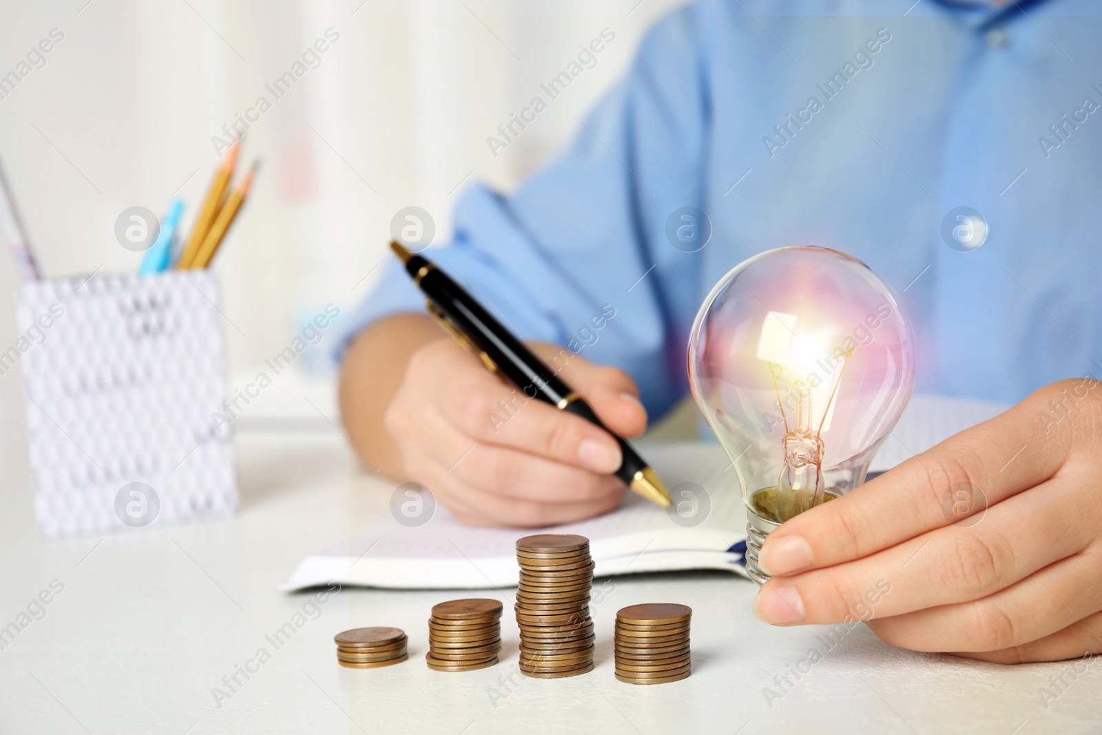 Woman with light bulb, notebook and coins at white table, closeup. Power saving Photo of Woman with light bulb, notebook and coins at white table, closeup. Power saving