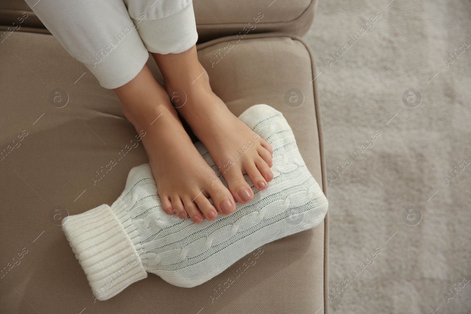 Woman warming feet with hot water bottle on sofa, closeup Photo of Woman warming feet with hot water bottle on sofa, closeup