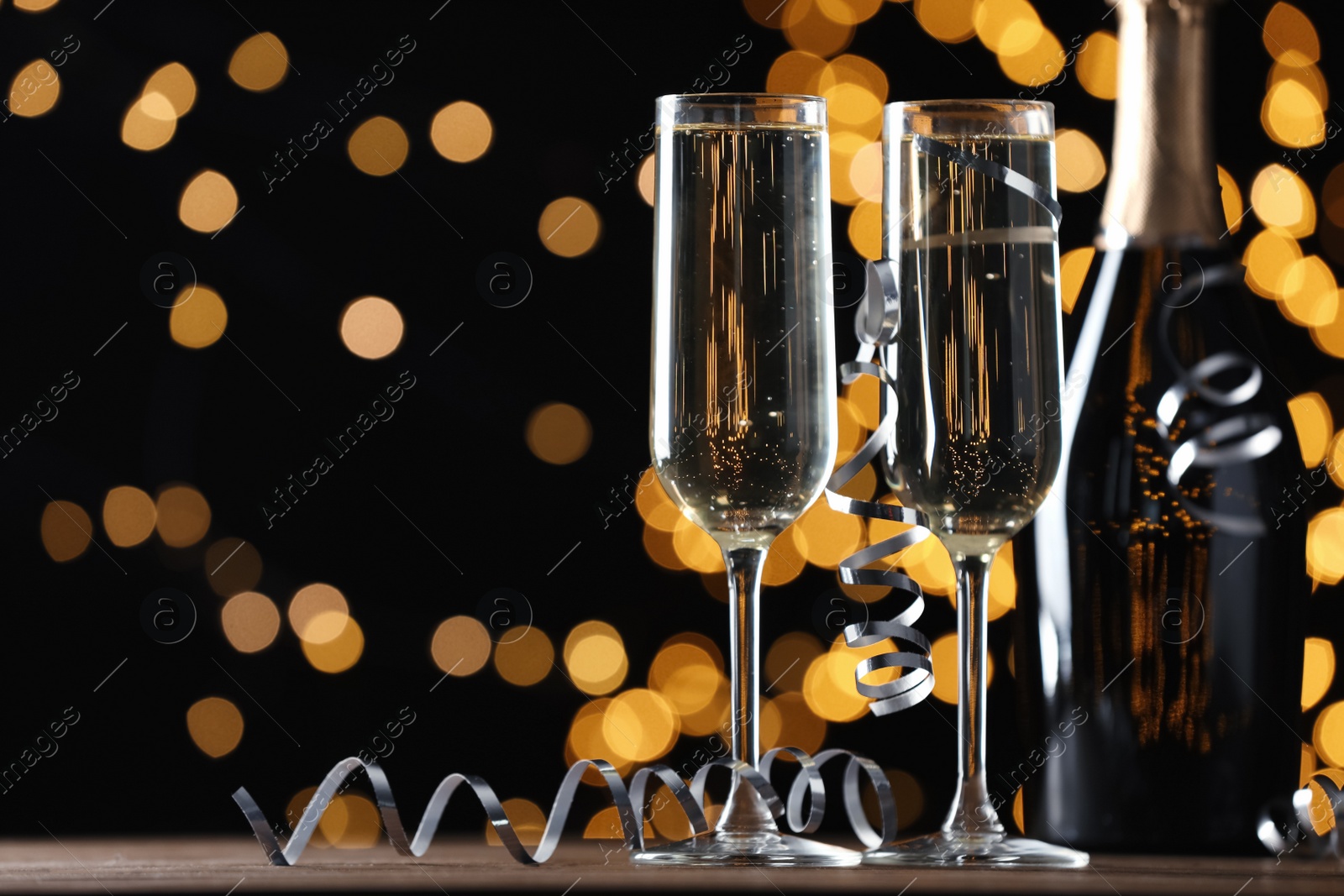 Photo of Glasses and bottle of champagne with serpentine streamers against blurred lights on black background. Space for text
