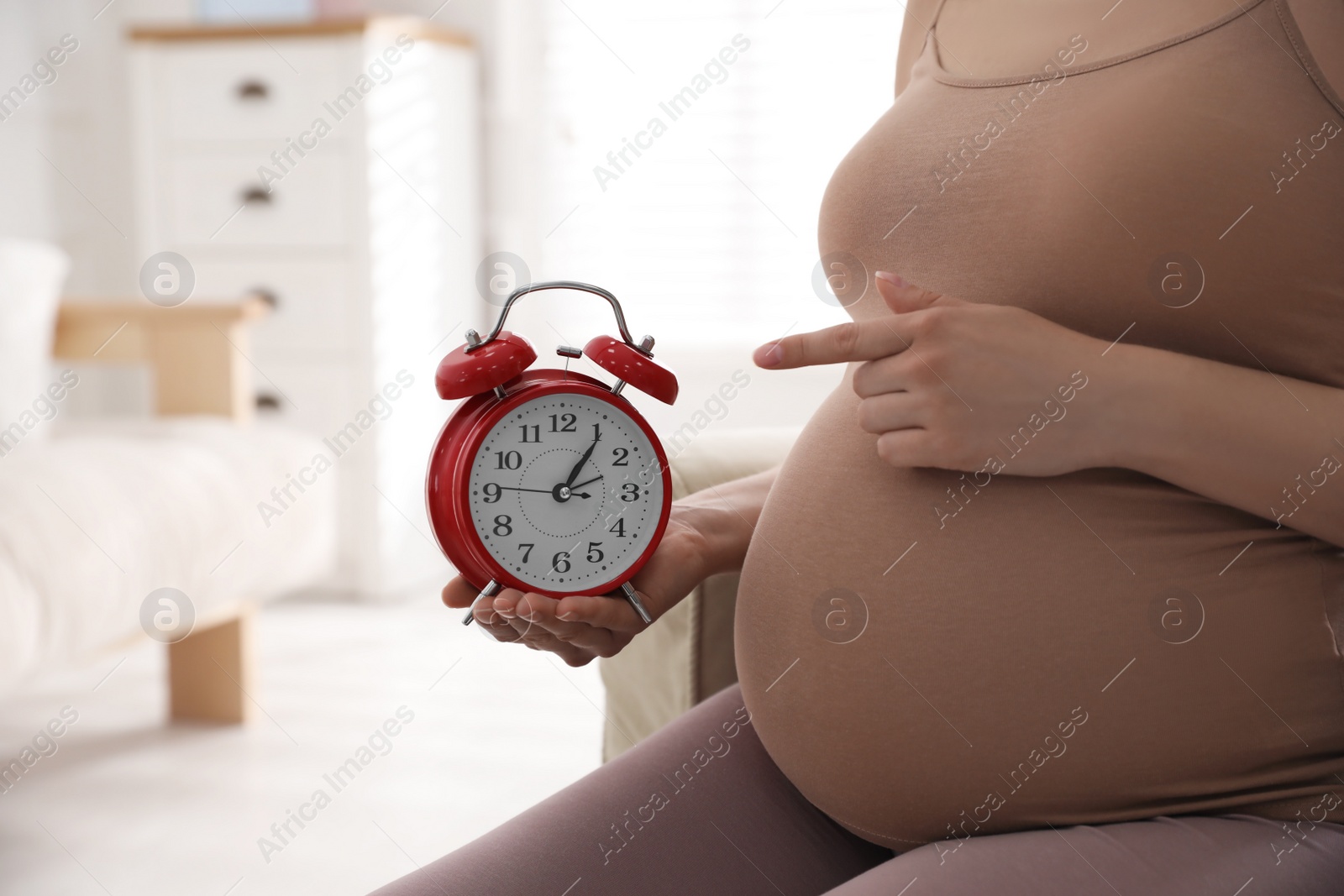 Young pregnant woman pointing at alarm clock near her belly indoors, closeup. Time to give birth Photo of Young pregnant woman pointing at alarm clock near her belly indoors, closeup. Time to give birth