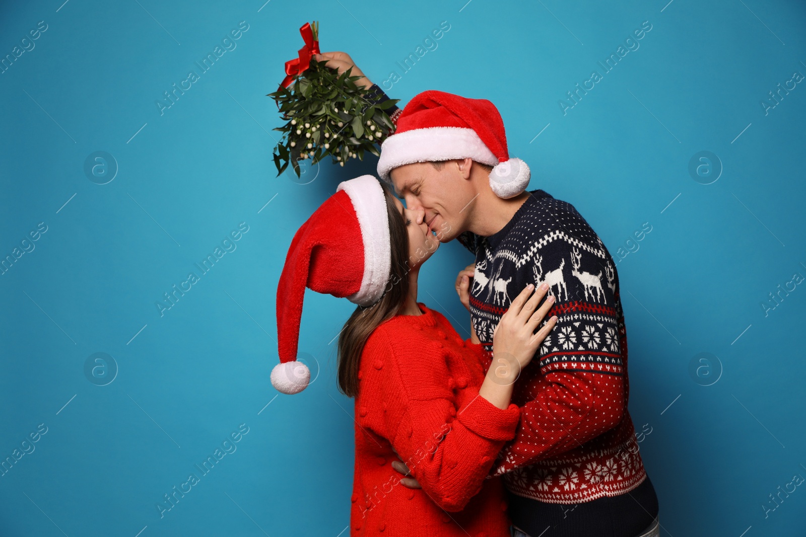 Photo of Happy couple kissing under mistletoe bunch on light blue background