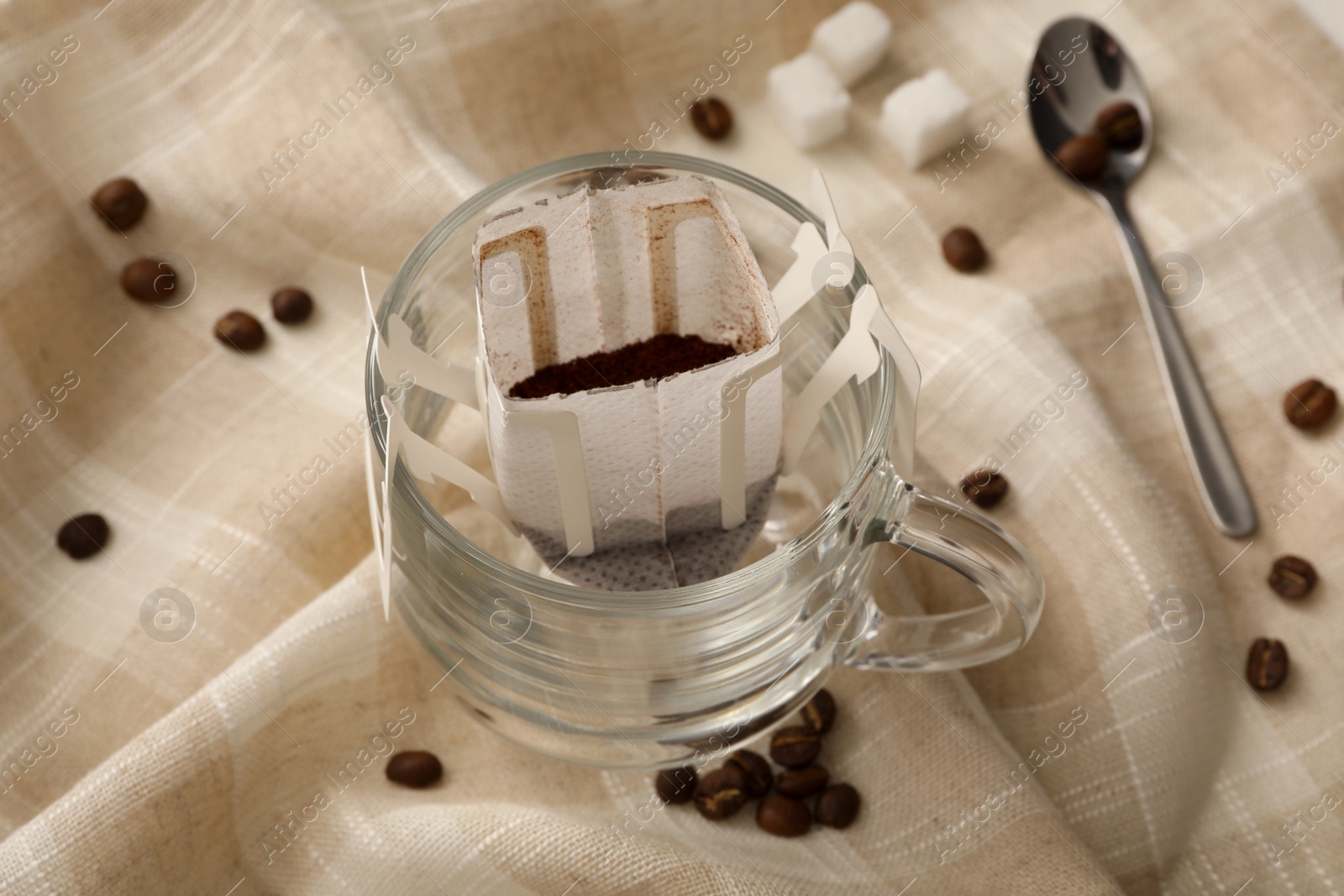 Glass cup with drip coffee bag and beans on beige fabric, closeup Photo of Glass cup with drip coffee bag and beans on beige fabric, closeup