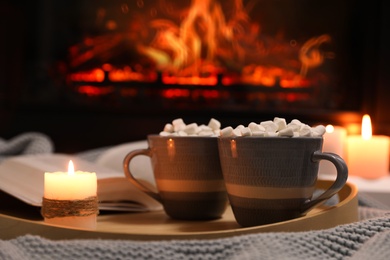 Cups of hot drink, candle and open book near fireplace indoors Photo of Cups of hot drink, candle and open book near fireplace indoors
