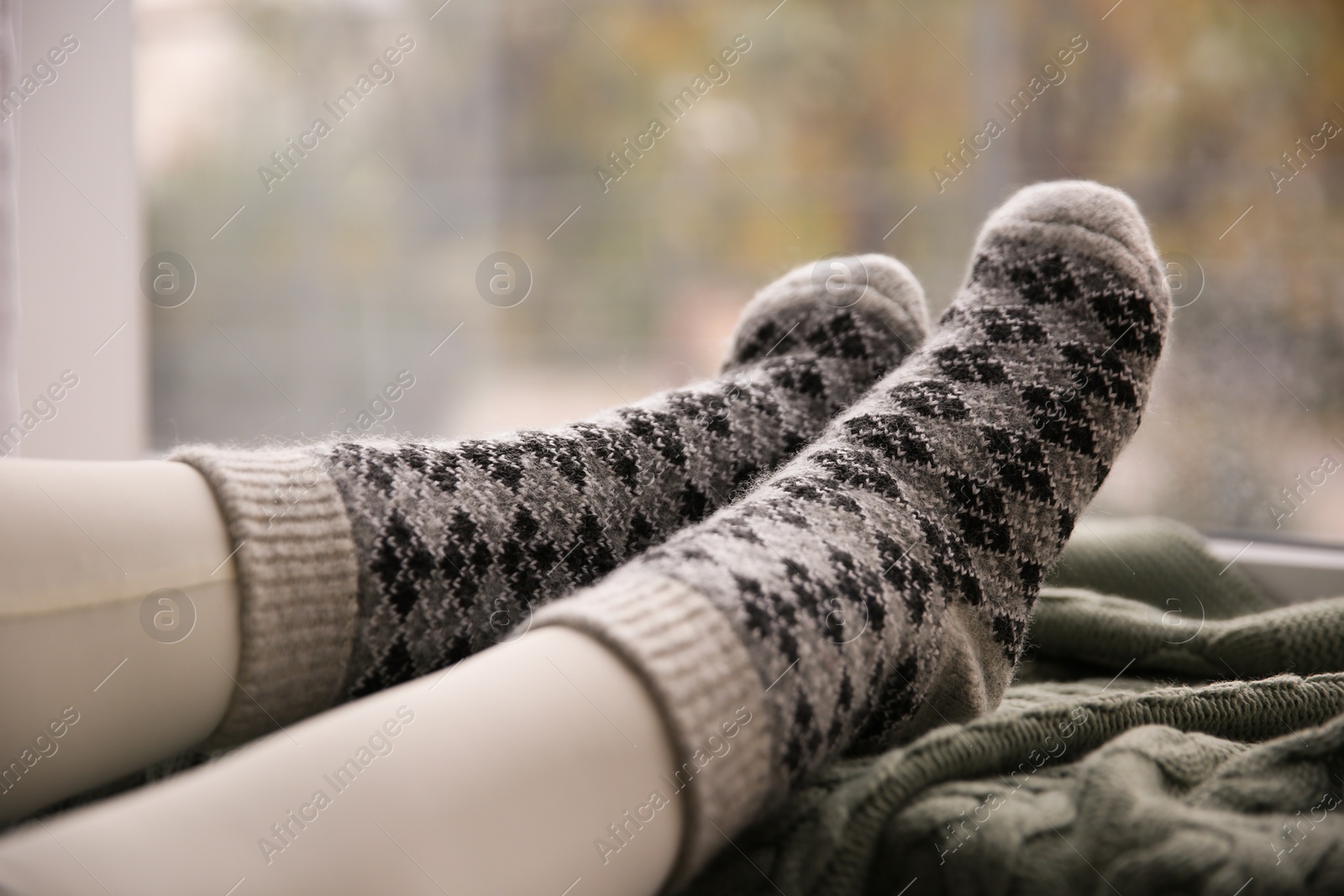 Woman wearing warm socks on knitted plaid near window, closeup. Cozy season Photo of Woman wearing warm socks on knitted plaid near window, closeup. Cozy season