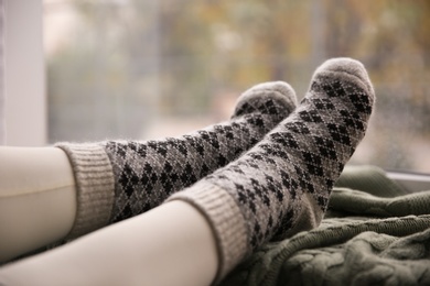 Woman wearing warm socks on knitted plaid near window, closeup. Cozy season Photo of Woman wearing warm socks on knitted plaid near window, closeup. Cozy season