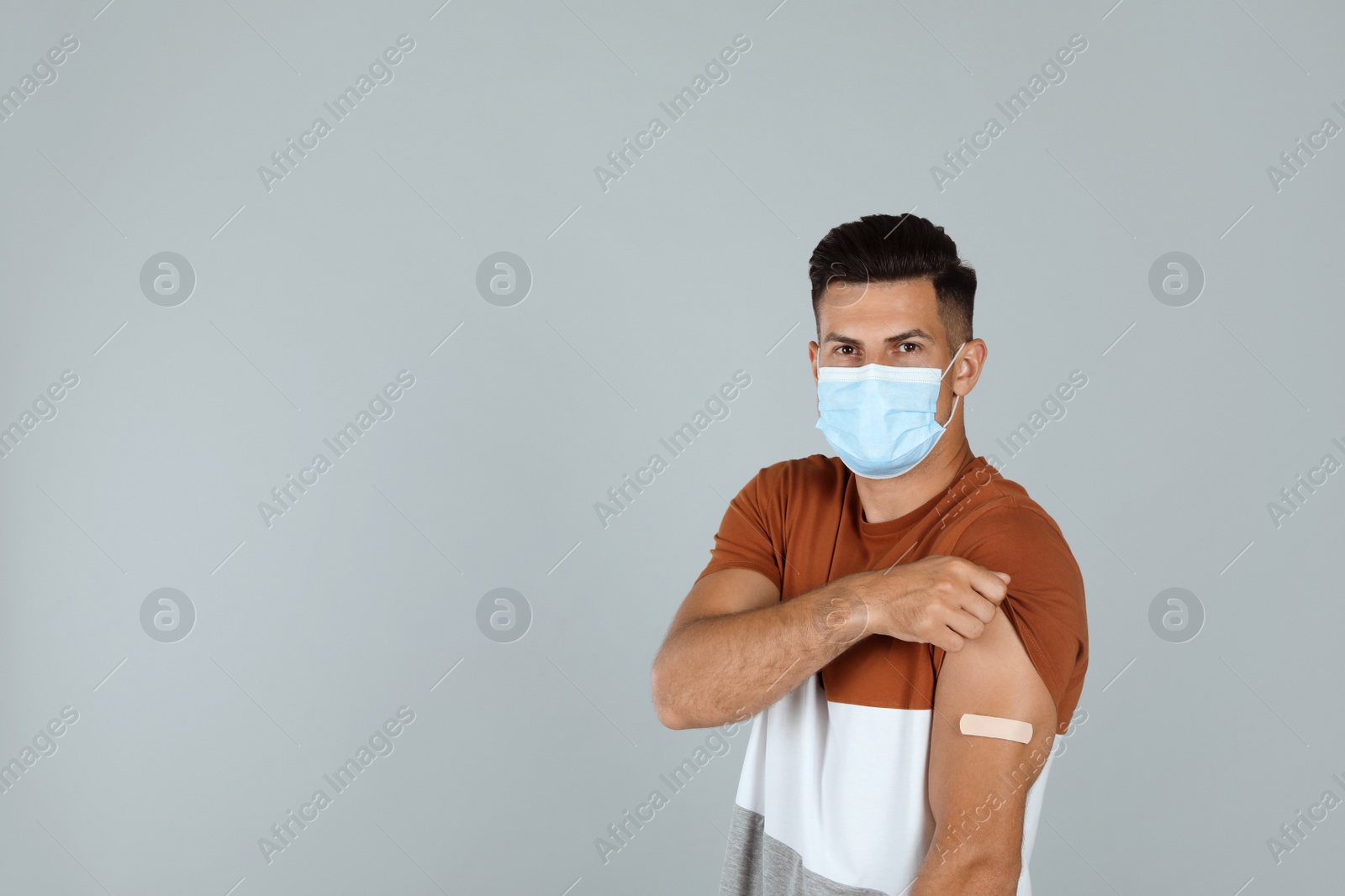 Vaccinated man with protective mask showing medical plaster on his arm against grey background. Space for text Photo of Vaccinated man with protective mask showing medical plaster on his arm against grey background. Space for text