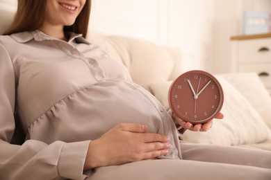 Young pregnant woman holding clock near her belly at home, closeup. Time to give birth Photo of Young pregnant woman holding clock near her belly at home, closeup. Time to give birth