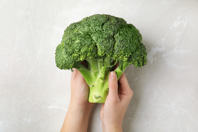 Photo of Woman holding fresh green broccoli over light grey marble table, top view