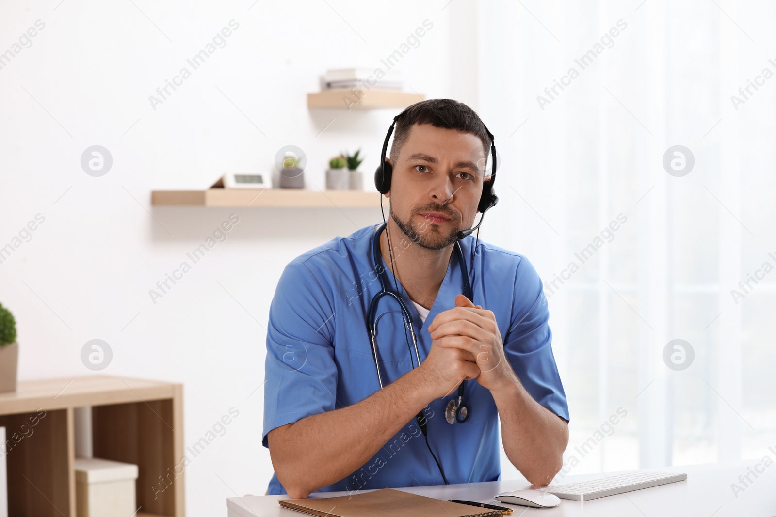Doctor with headset sitting at desk in clinic. Health service hotline Photo of Doctor with headset sitting at desk in clinic. Health service hotline