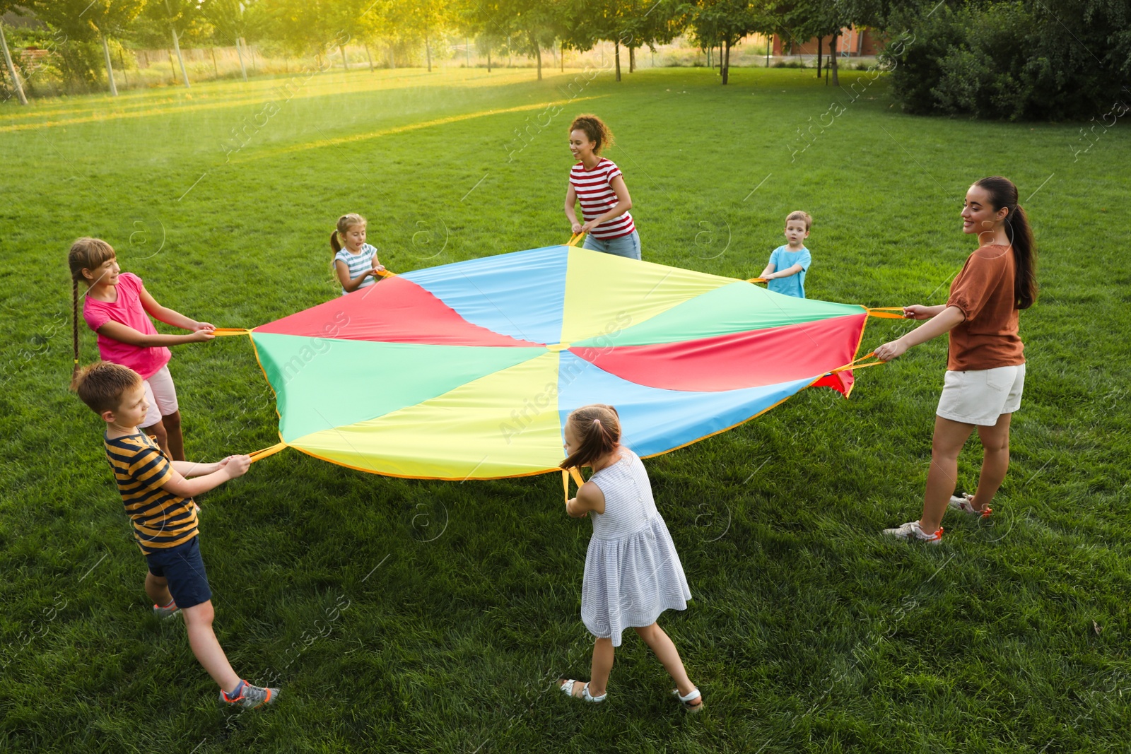 Group of children and teachers playing with rainbow playground parachute on green grass. Summer camp activity Photo of Group of children and teachers playing with rainbow playground parachute on green grass. Summer camp activity