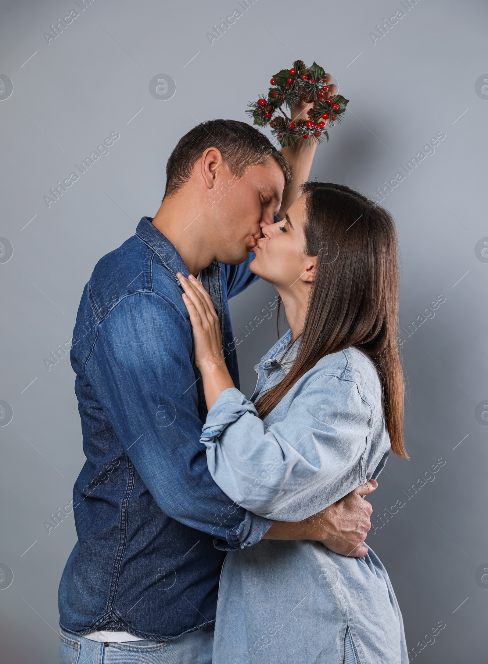 Happy couple kissing under mistletoe wreath on grey background Photo of Happy couple kissing under mistletoe wreath on grey background