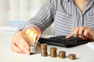 Woman with light bulb, calculator and coins at white table, closeup. Power saving Photo of Woman with light bulb, calculator and coins at white table, closeup. Power saving