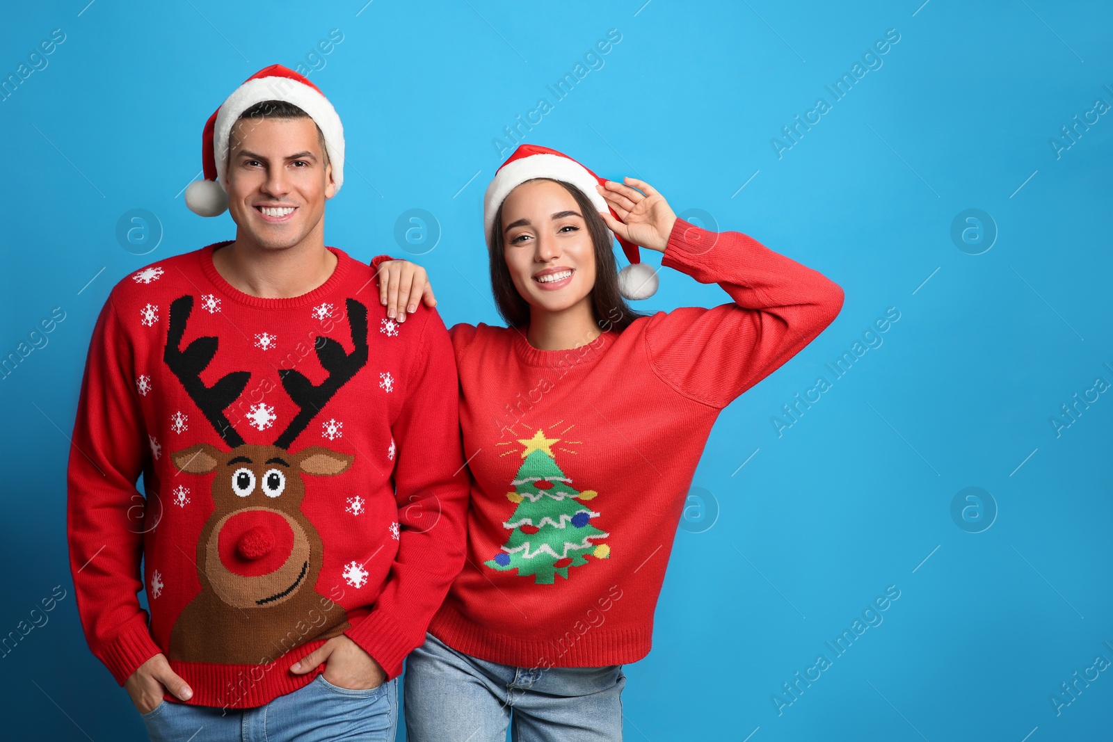 Couple in Christmas sweaters and Santa hats on blue background, space for text Photo of Couple in Christmas sweaters and Santa hats on blue background, space for text