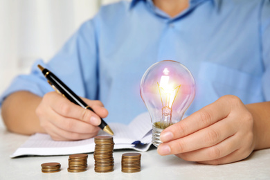 Woman with light bulb, notebook and coins at white table, closeup. Power saving Photo of Woman with light bulb, notebook and coins at white table, closeup. Power saving