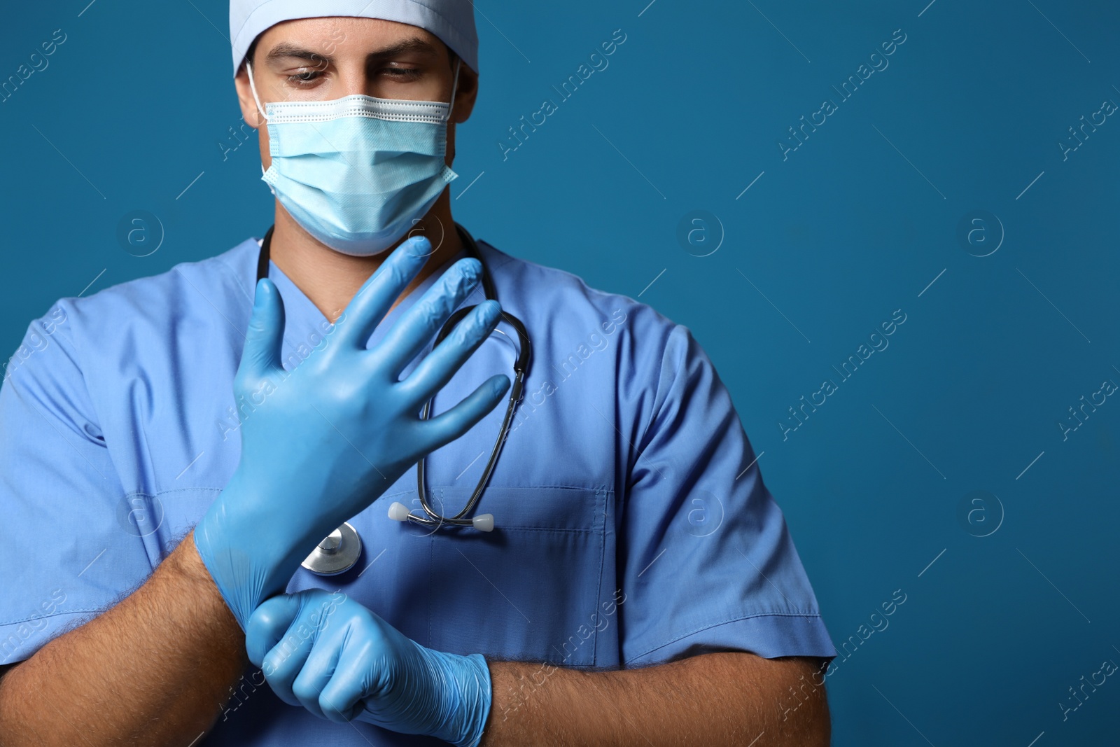 Doctor in protective mask putting on medical gloves against blue background Photo of Doctor in protective mask putting on medical gloves against blue background