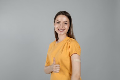 Vaccinated woman with medical plaster on her arm showing thumb up against grey background Photo of Vaccinated woman with medical plaster on her arm showing thumb up against grey background