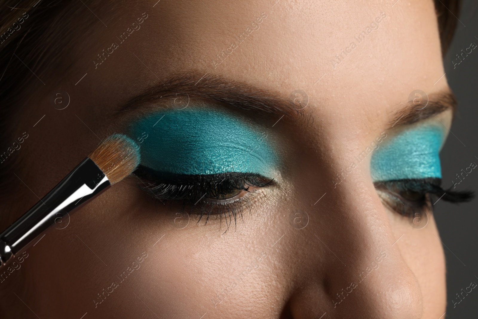 Applying cyan eye shadow onto woman's face on grey background, closeup. Beautiful evening makeup Photo of Applying cyan eye shadow onto woman's face on grey background, closeup. Beautiful evening makeup