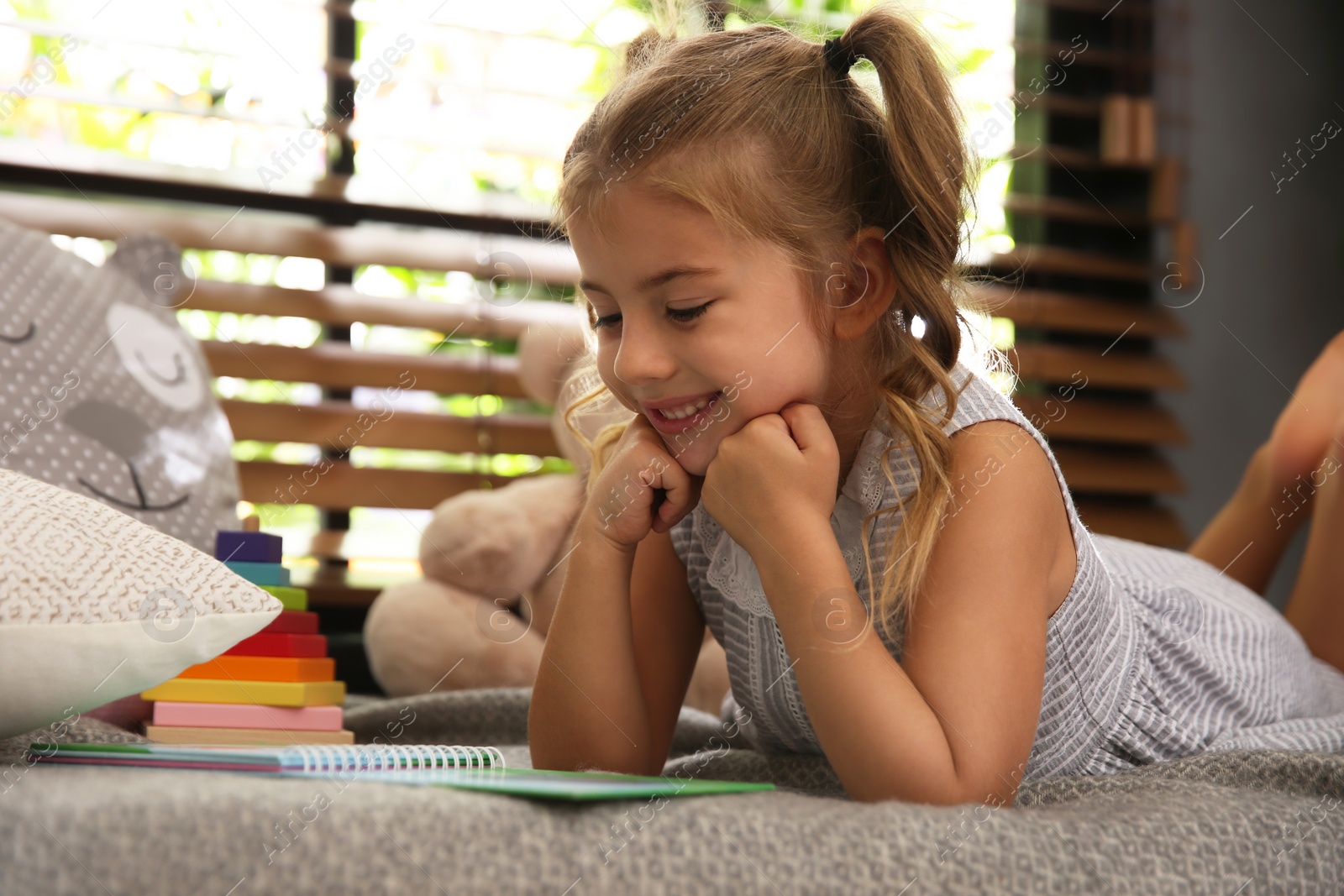 Photo of Cute little girl reading book near window at home