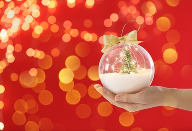Woman holding transparent Christmas ornament with small tree, fairy lights and snow against red background, bokeh effect. Space for text Image of Woman holding transparent Christmas ornament with small tree, fairy lights and snow against red background, bokeh effect. Space for text