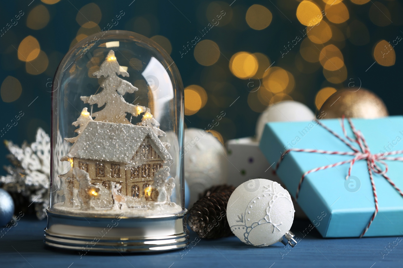 Beautiful snow globe, gift box and Christmas decor on blue table against blurred festive lights Photo of Beautiful snow globe, gift box and Christmas decor on blue table against blurred festive lights