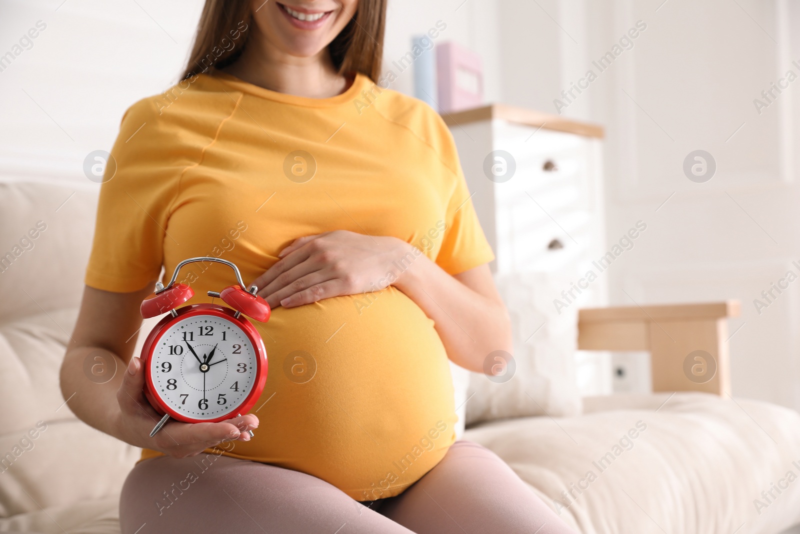 Young pregnant woman holding alarm clock near her belly at home, closeup. Time to give birth Photo of Young pregnant woman holding alarm clock near her belly at home, closeup. Time to give birth