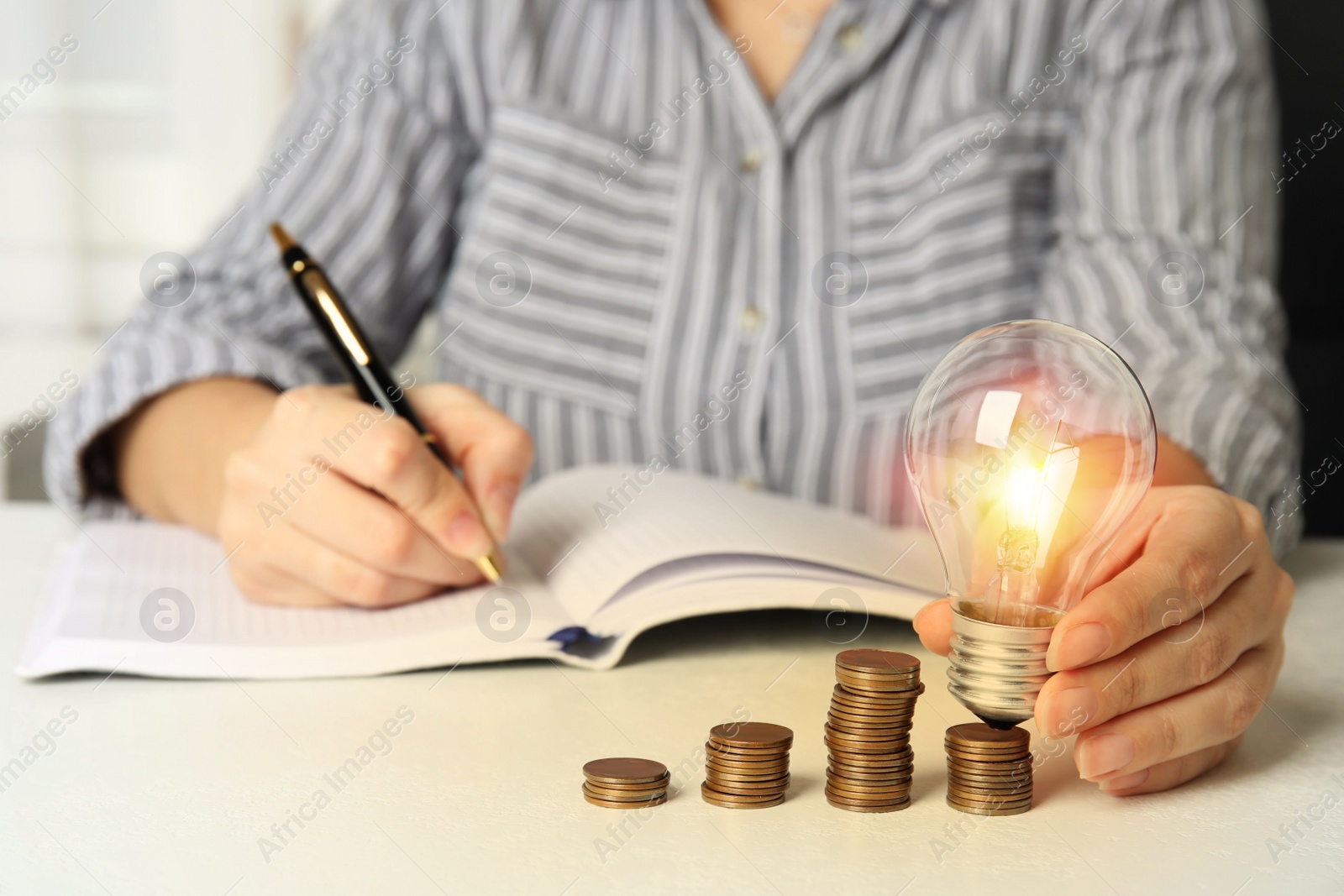 Woman with light bulb, notebook and coins at white table, closeup. Power saving Photo of Woman with light bulb, notebook and coins at white table, closeup. Power saving