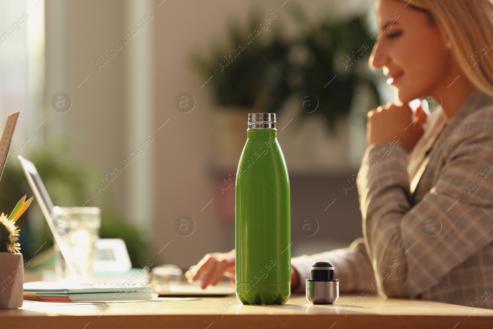 Woman working at table in modern office, focus on green thermos bottle Photo of Woman working at table in modern office, focus on green thermos bottle