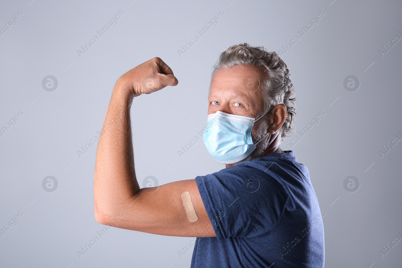 Senior man in protective mask showing arm with bandage after vaccination on grey background Photo of Senior man in protective mask showing arm with bandage after vaccination on grey background