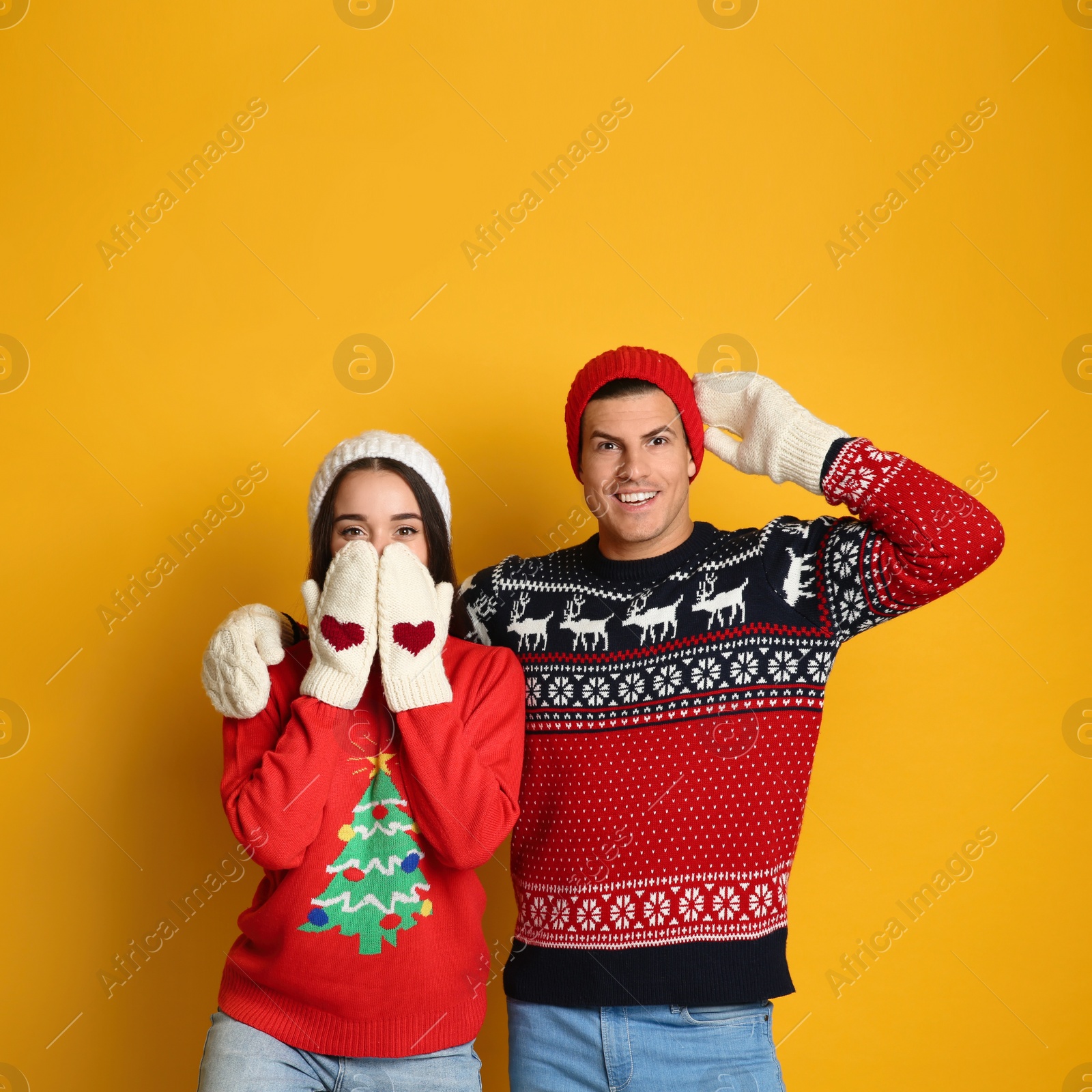 Couple in Christmas sweaters, knitted mittens and hats on yellow background Photo of Couple in Christmas sweaters, knitted mittens and hats on yellow background
