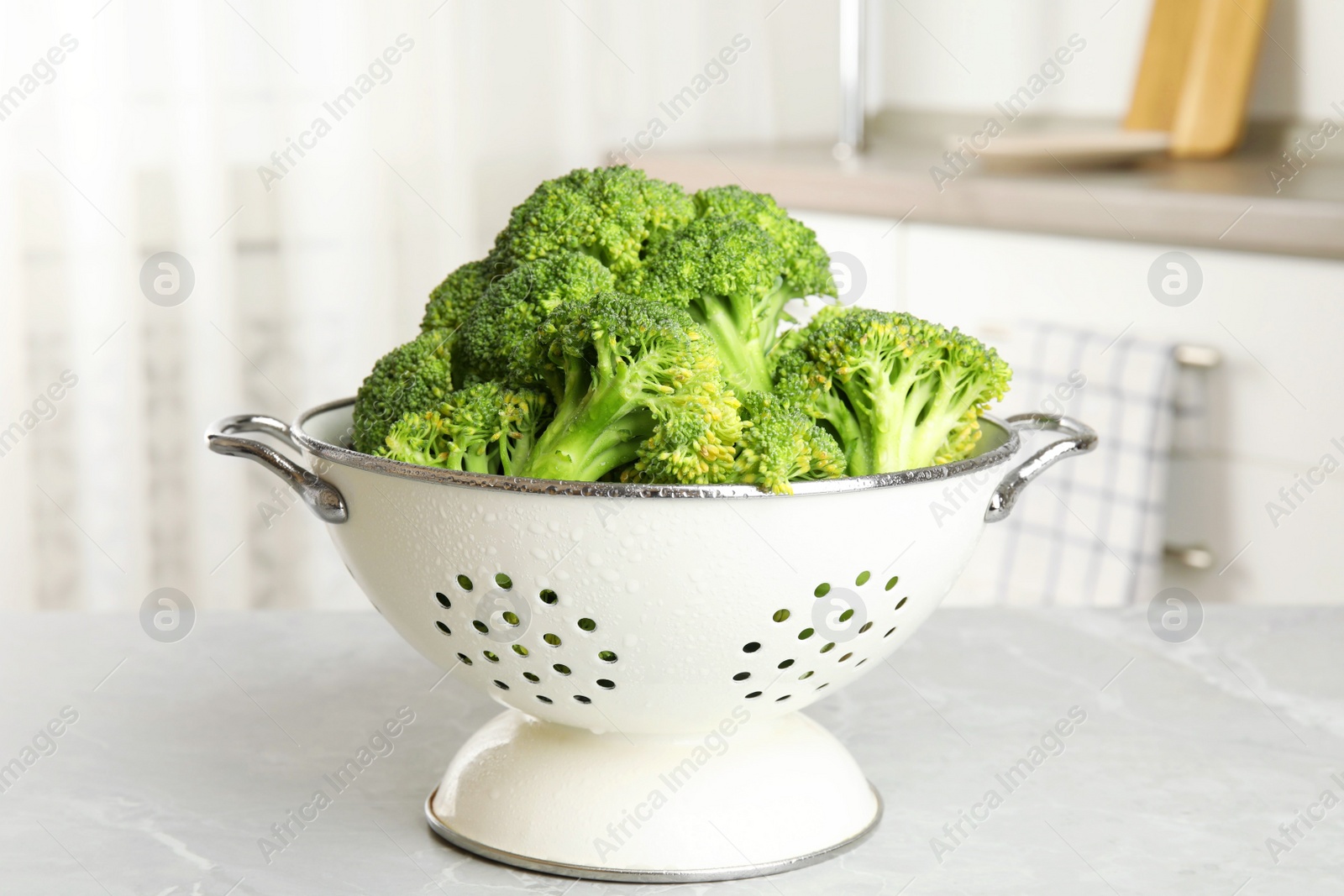 Raw green broccoli in colander on light grey marble table indoors Photo of Raw green broccoli in colander on light grey marble table indoors