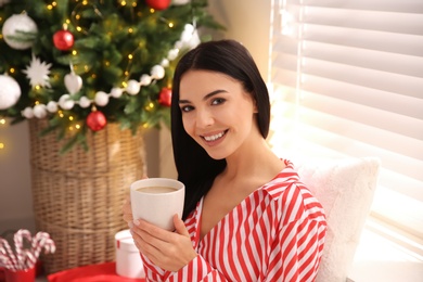 Young woman with cup of coffee near Christmas tree at home Photo of Young woman with cup of coffee near Christmas tree at home