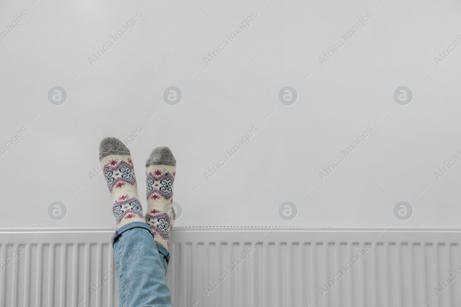 Woman warming legs on heating radiator near white wall, closeup. Space for text Photo of Woman warming legs on heating radiator near white wall, closeup. Space for text