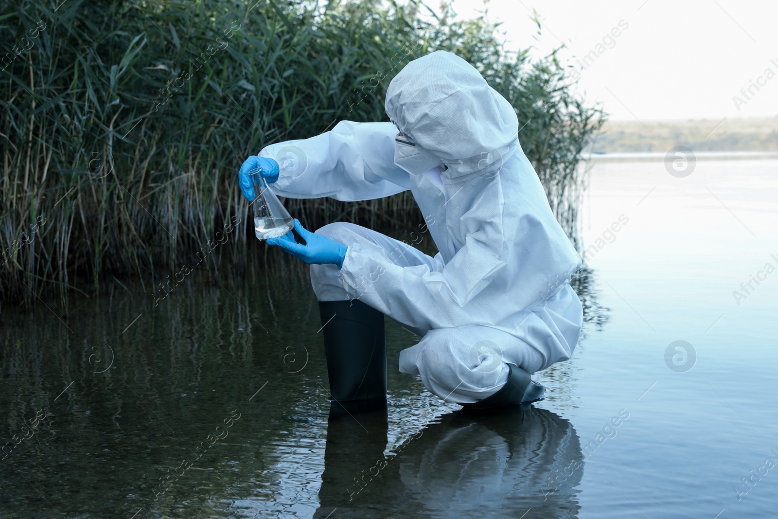 Scientist in chemical protective suit with conical flask taking sample from river for analysis Photo of Scientist in chemical protective suit with conical flask taking sample from river for analysis