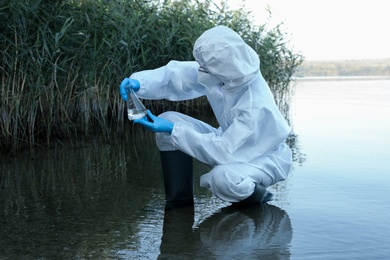 Scientist in chemical protective suit with conical flask taking sample from river for analysis Photo of Scientist in chemical protective suit with conical flask taking sample from river for analysis