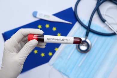 Doctor holding sample tube with label Coronavirus Test above medical items and European Union flag, closeup Photo of Doctor holding sample tube with label Coronavirus Test above medical items and European Union flag, closeup