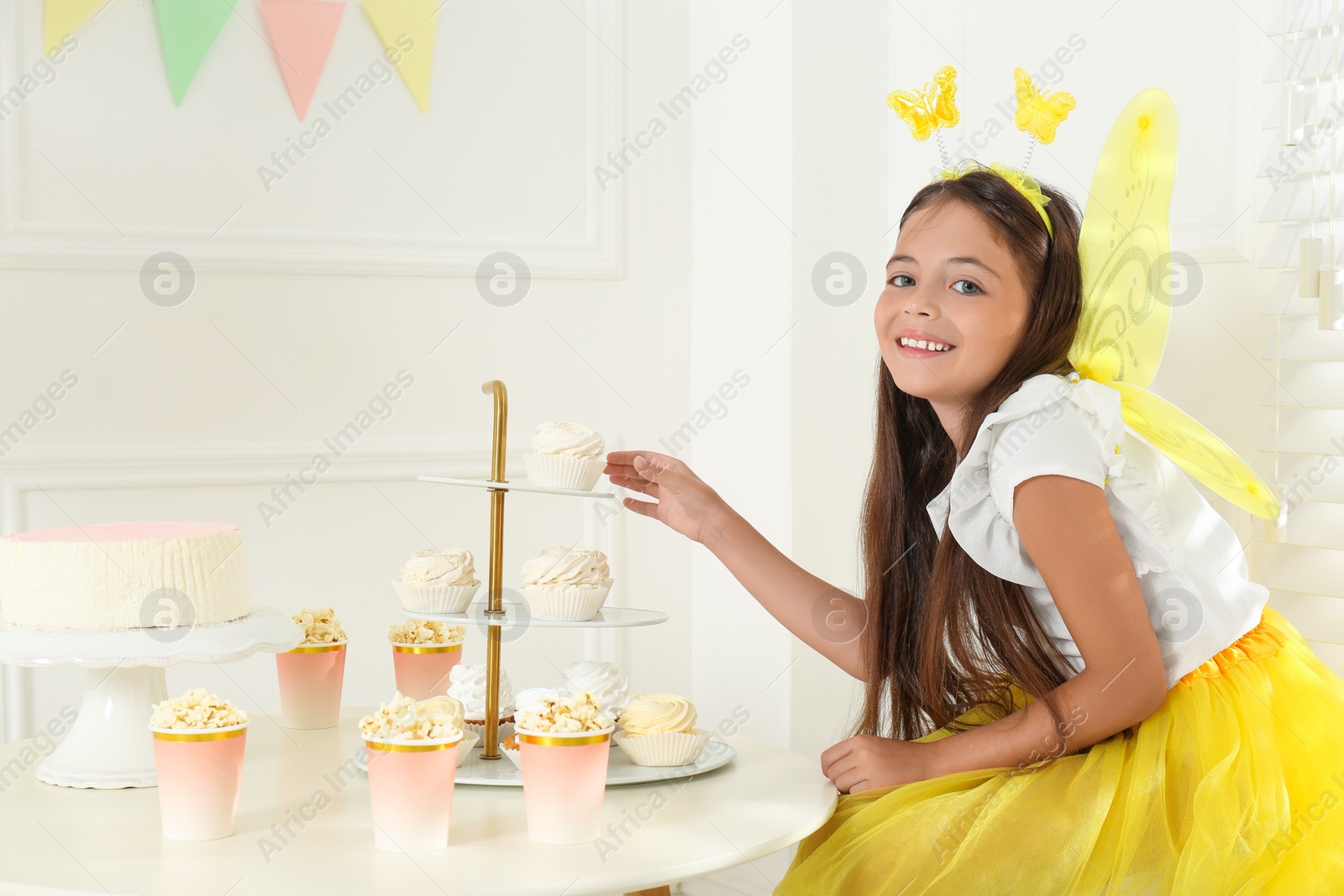 Cute little girl wearing fairy costume at table with desserts in decorated room Photo of Cute little girl wearing fairy costume at table with desserts in decorated room