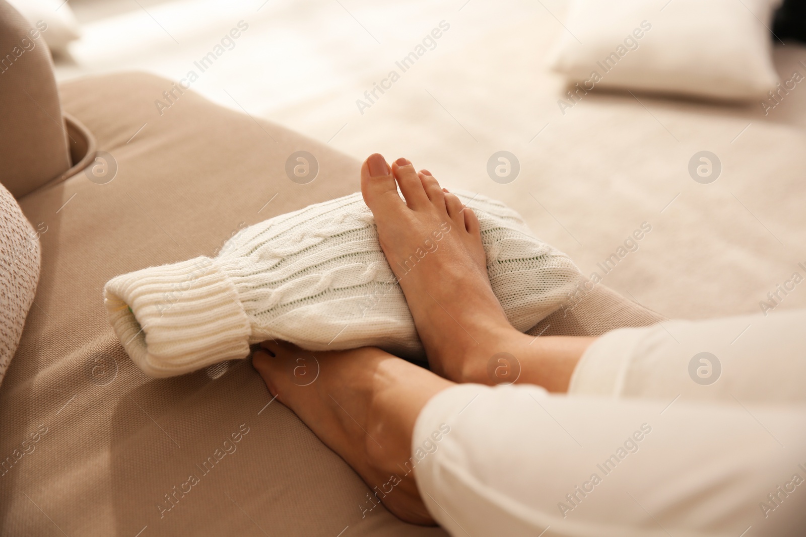 Woman warming feet with hot water bottle on sofa, closeup Photo of Woman warming feet with hot water bottle on sofa, closeup