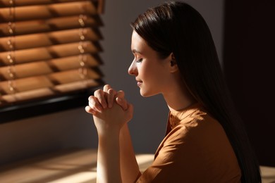 Religious young woman with clasped hands praying indoors Photo of Religious young woman with clasped hands praying indoors